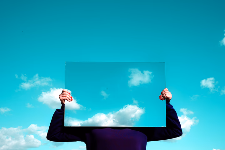 A person holds a rectangular mirror in front of their head, reflecting the blue sky and clouds, blending the mirror with the background.