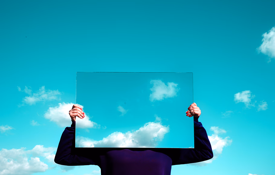 A person holds a rectangular mirror in front of their head, reflecting the blue sky and clouds, blending the mirror with the background.